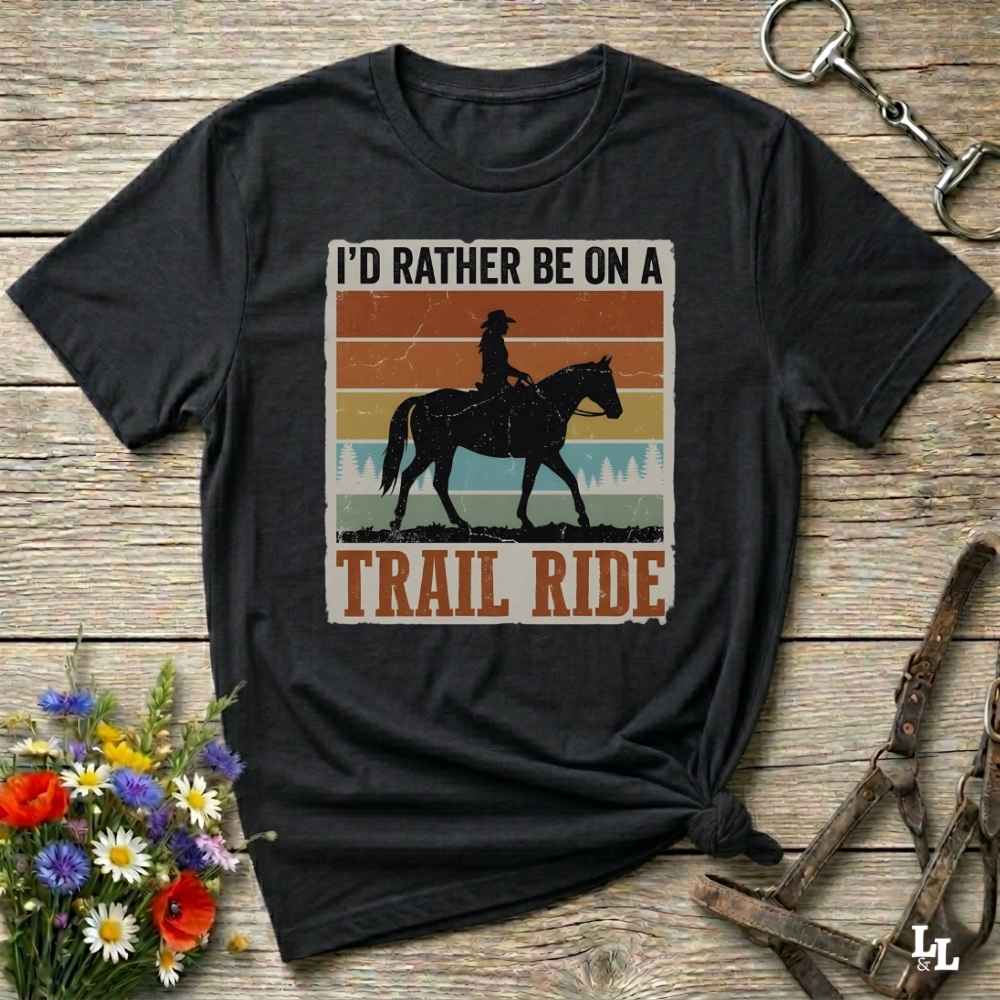 A black graphic t-shirt featuring a western-themed 'Trail Ride' design with a silhouette of a cowboy on a horse against retro sunset stripes, displayed on a rustic wooden background with equestrian accessories.