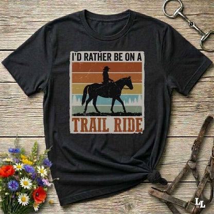 A black graphic t-shirt featuring a western-themed 'Trail Ride' design with a silhouette of a cowboy on a horse against retro sunset stripes, displayed on a rustic wooden background with equestrian accessories.