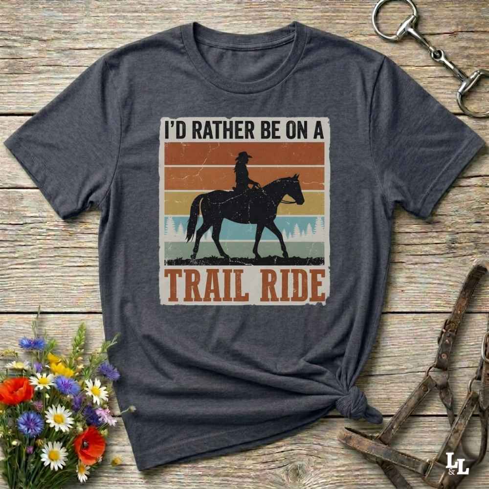 A dark heather gray graphic t-shirt featuring a retro-style western design with a cowboy on a horse and the text 'I'D RATHER BE ON A TRAIL RIDE'. The shirt is styled in a flat-lay on a rustic wooden background with equestrian accessories and flowers.