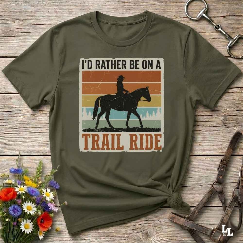 An olive green short-sleeve t-shirt with a retro-style graphic of a cowboy riding a horse and the text 'I'D RATHER BE ON A TRAIL RIDE'. The shirt is displayed flat on a rustic wooden background with decorative floral and leather props.