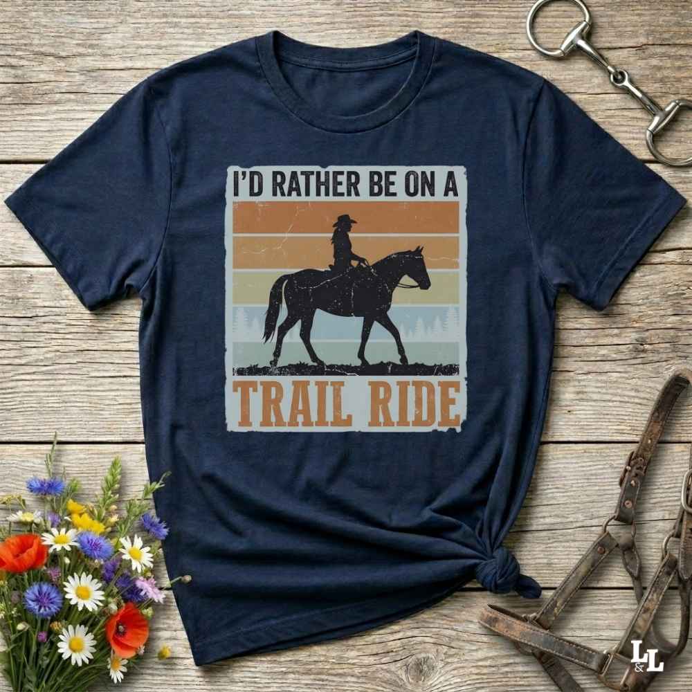 A navy blue graphic t-shirt laid flat on a rustic wooden surface, styled with equestrian accessories and a small bouquet of wildflowers. The shirt features a retro-style graphic with the text 'I'D RATHER BE ON A TRAIL RIDE' and a silhouette of a cowboy riding a horse against a striped sunset background.