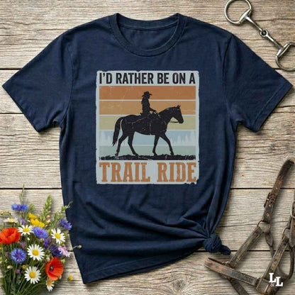 A navy blue graphic t-shirt laid flat on a rustic wooden surface, styled with equestrian accessories and a small bouquet of wildflowers. The shirt features a retro-style graphic with the text 'I'D RATHER BE ON A TRAIL RIDE' and a silhouette of a cowboy riding a horse against a striped sunset background.