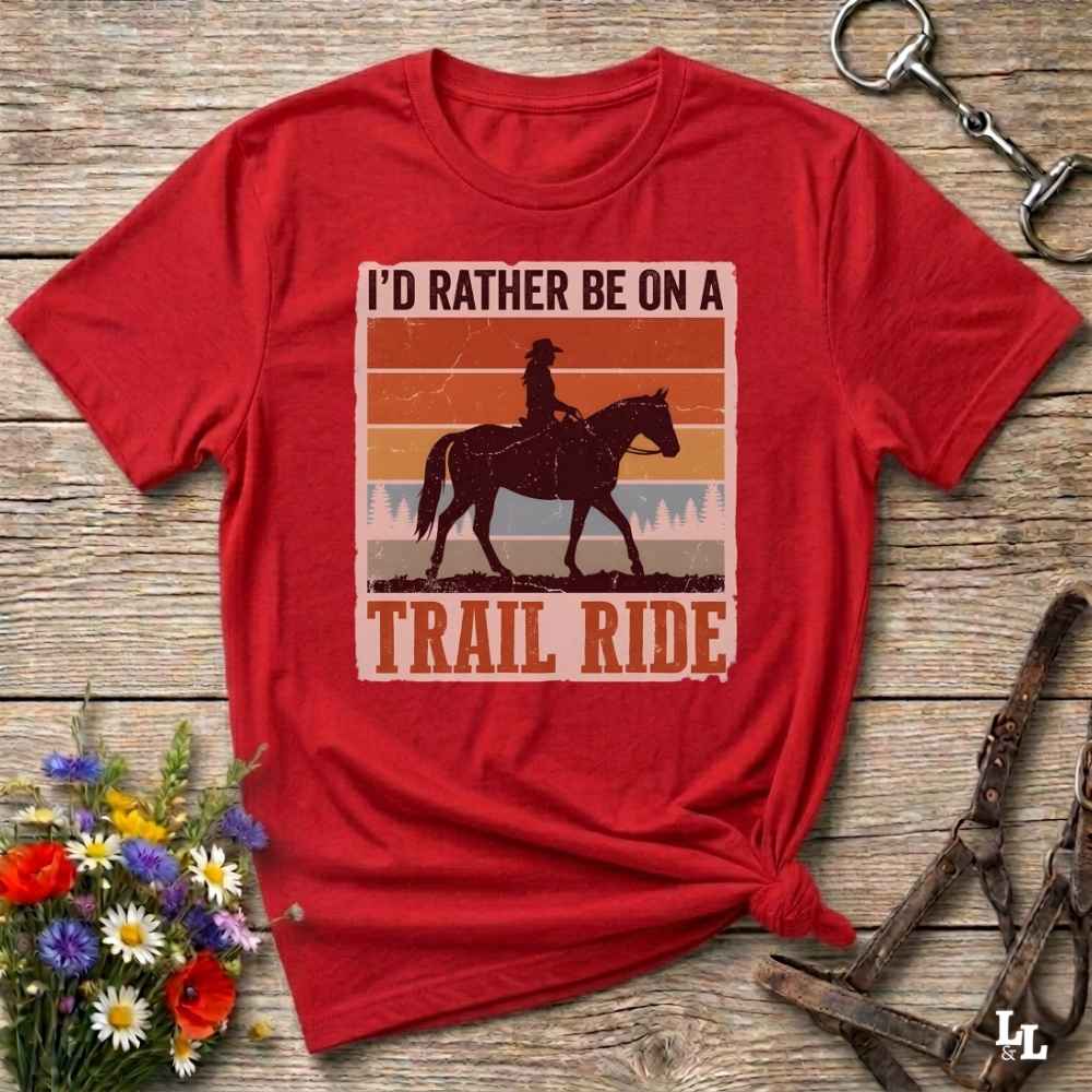 A red short-sleeve graphic T-shirt laid flat on a rustic wood surface, with a distressed rectangular print showing a cowboy riding a horse and the text “I'D RATHER BE ON A TRAIL RIDE.” Decorative flowers and tack are placed around it as props.