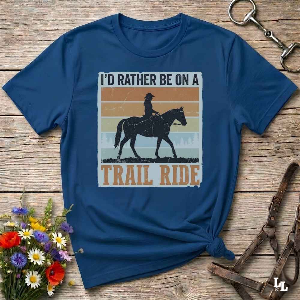A navy blue graphic t-shirt laid flat on a rustic wooden surface, styled with a small bouquet of flowers and equestrian tack. The shirt features a retro-style graphic with a cowboy riding a horse and the text 'I'D RATHER BE ON A TRAIL RIDE'.