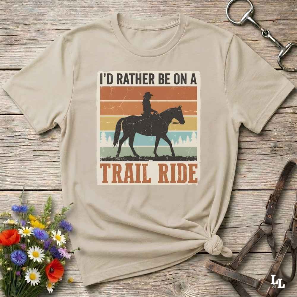 A beige graphic t-shirt featuring a retro western design with a cowboy riding a horse and the text 'I'D RATHER BE ON A TRAIL RIDE'. The shirt is styled in a flat-lay on a rustic wooden background with equestrian accessories and a small bouquet of wildflowers.