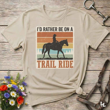 A beige graphic t-shirt featuring a retro western design with a cowboy riding a horse and the text 'I'D RATHER BE ON A TRAIL RIDE'. The shirt is styled in a flat-lay on a rustic wooden background with equestrian accessories and a small bouquet of wildflowers.