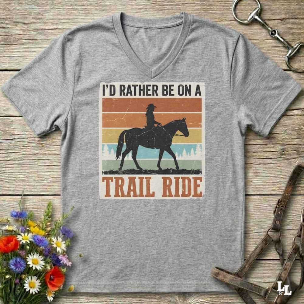 A heather gray V-neck t-shirt laid flat on a rustic wooden surface, featuring a retro-style graphic of a cowboy riding a horse with the text 'I'D RATHER BE ON A TRAIL RIDE'. The shirt is styled with equestrian props, including a halter, a horse bit, and a small bouquet of wildflowers.