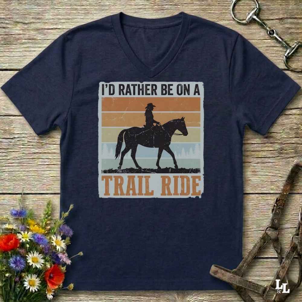 A navy blue V-neck t-shirt laid flat on a rustic wooden surface, featuring a retro-style graphic of a cowboy riding a horse with the text 'I'D RATHER BE ON A TRAIL RIDE'. The shirt is styled with equestrian props, including a halter, a reining rope, and a small bouquet of wildflowers.