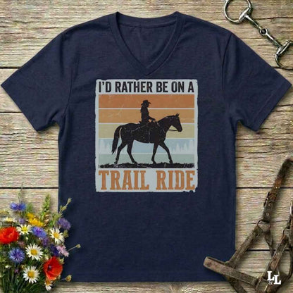 A navy blue V-neck t-shirt laid flat on a rustic wooden surface, featuring a retro-style graphic of a cowboy riding a horse with the text 'I'D RATHER BE ON A TRAIL RIDE'. The shirt is styled with equestrian props, including a halter, a reining rope, and a small bouquet of wildflowers.