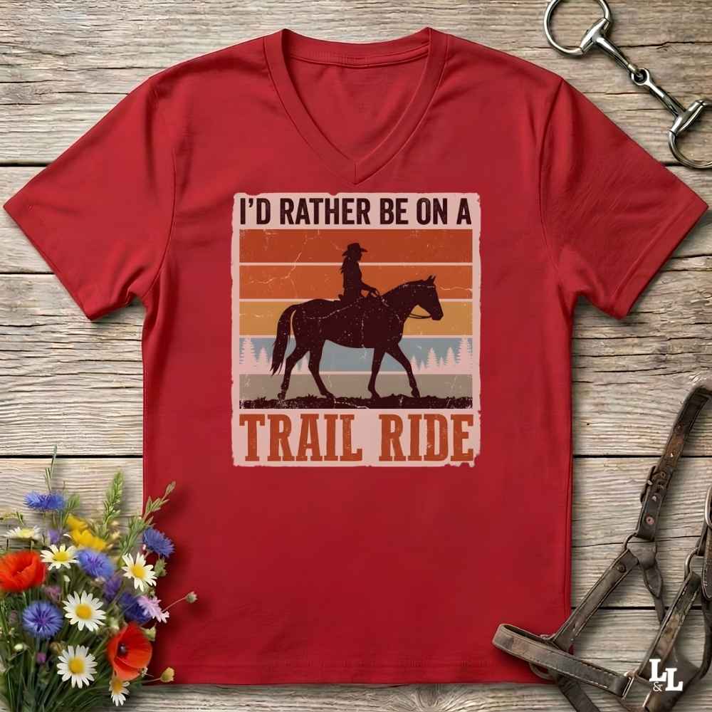 A red V-neck t-shirt laid flat on a rustic wooden surface, featuring a retro western graphic of a cowboy riding a horse with the text 'I'D RATHER BE ON A TRAIL RIDE'. The shirt is styled with equestrian tack and a small bouquet of wildflowers.