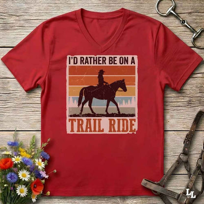 A red V-neck t-shirt laid flat on a rustic wooden surface, featuring a retro western graphic of a cowboy riding a horse with the text 'I'D RATHER BE ON A TRAIL RIDE'. The shirt is styled with equestrian tack and a small bouquet of wildflowers.