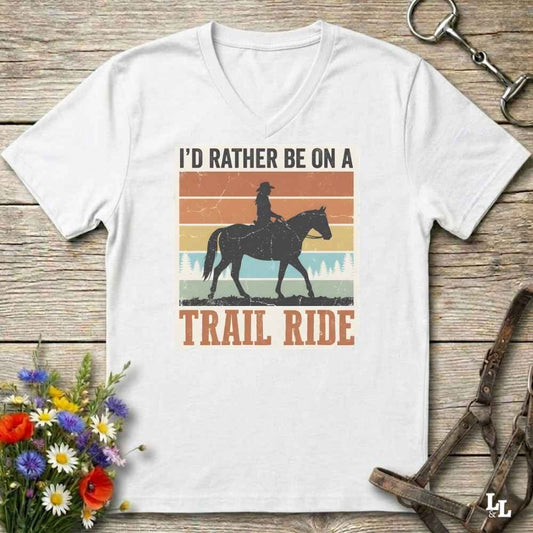 A white V-neck t-shirt with a retro western graphic of a cowboy riding a horse, with the text 'I'D RATHER BE ON A TRAIL RIDE'. The shirt is displayed flat on a rustic wooden background with equestrian accessories and flowers.