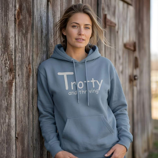 Woman wearing a blue hoodie with 'Trotty and thriving' text against a wooden barn. Trotty and Thriving Hoodie for Horse Lovers, Trail Riding Hoodie Equestrian Wear, perfect gift for horse lovers, mothers day gift, fathers day gift