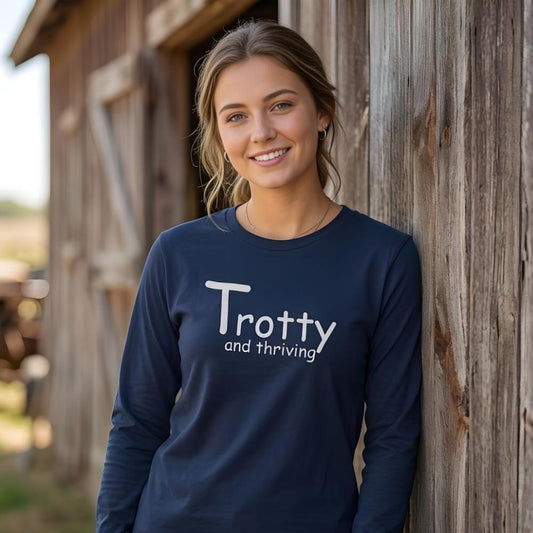 Woman wearing a navy blue shirt with 'Trotty and thriving' text, leaning against a wooden barn. Trotty and Thriving Long Sleeve Tee for Horse Riders, Trail Riding Tee, perfect gift for horse lovers, mothers day gift, father days gift