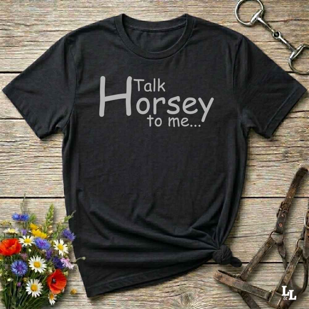 A black short-sleeve graphic T-shirt laid flat on a rustic wooden surface, with white text reading 'Talk Horsey to me.