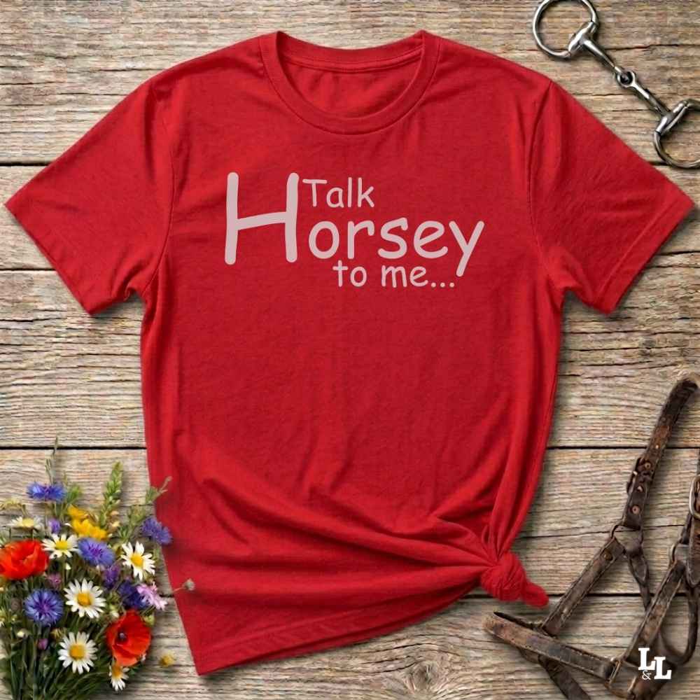 A red short-sleeve graphic T-shirt laid flat on a rustic wooden surface, with white text reading 'Talk Horsey to me.
