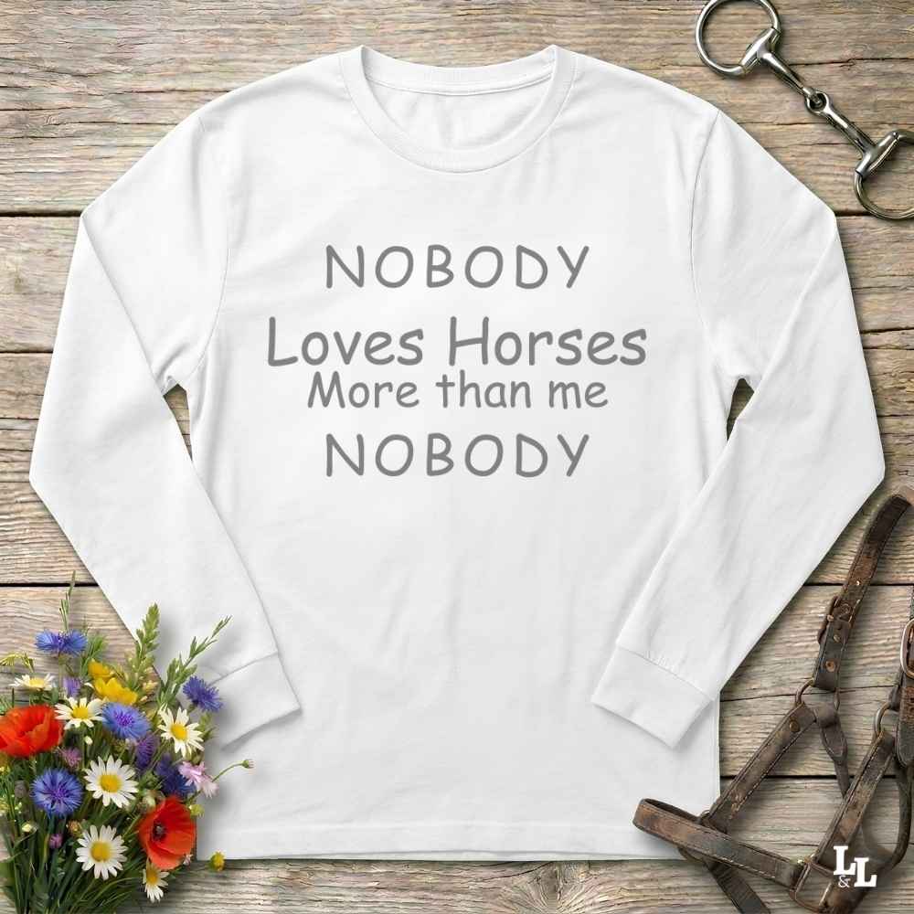 A white long-sleeve t-shirt with gray text reading 'NOBODY Loves Horses More than me NOBODY' laid flat on a rustic wooden background with decorative horse tack and flowers.