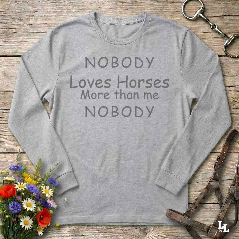 A light gray long-sleeve t-shirt with the text 'NOBODY Loves Horses More than me NOBODY' printed in dark gray. The shirt is displayed in a flat-lay arrangement on a rustic wooden background, surrounded by equestrian-themed props including a horse bit, leather bridle, and a small bouquet of colorful wildflowers.
