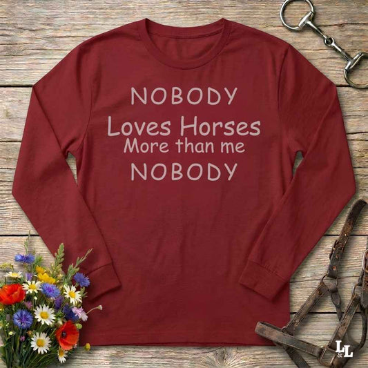 A long-sleeve crewneck t-shirt in maroon red with the text 'NOBODY Loves Horses More than me NOBODY' printed in white across the chest. The shirt is displayed flat on a rustic wooden background, styled with equestrian leather tack, a horse bit, and a small bouquet of colorful wildflowers.