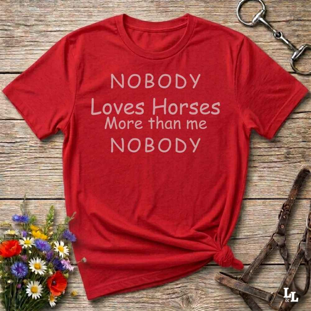 A red short-sleeve graphic T-shirt laid flat on a rustic wooden surface, with white text reading 'NOBODY Loves Horses More than me NOBODY.' Decorative flowers and horse tack are placed around it as styling props.