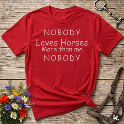 A red short-sleeve graphic T-shirt laid flat on a rustic wooden surface, with white text reading 'NOBODY Loves Horses More than me NOBODY.' Decorative flowers and horse tack are placed around it as styling props.