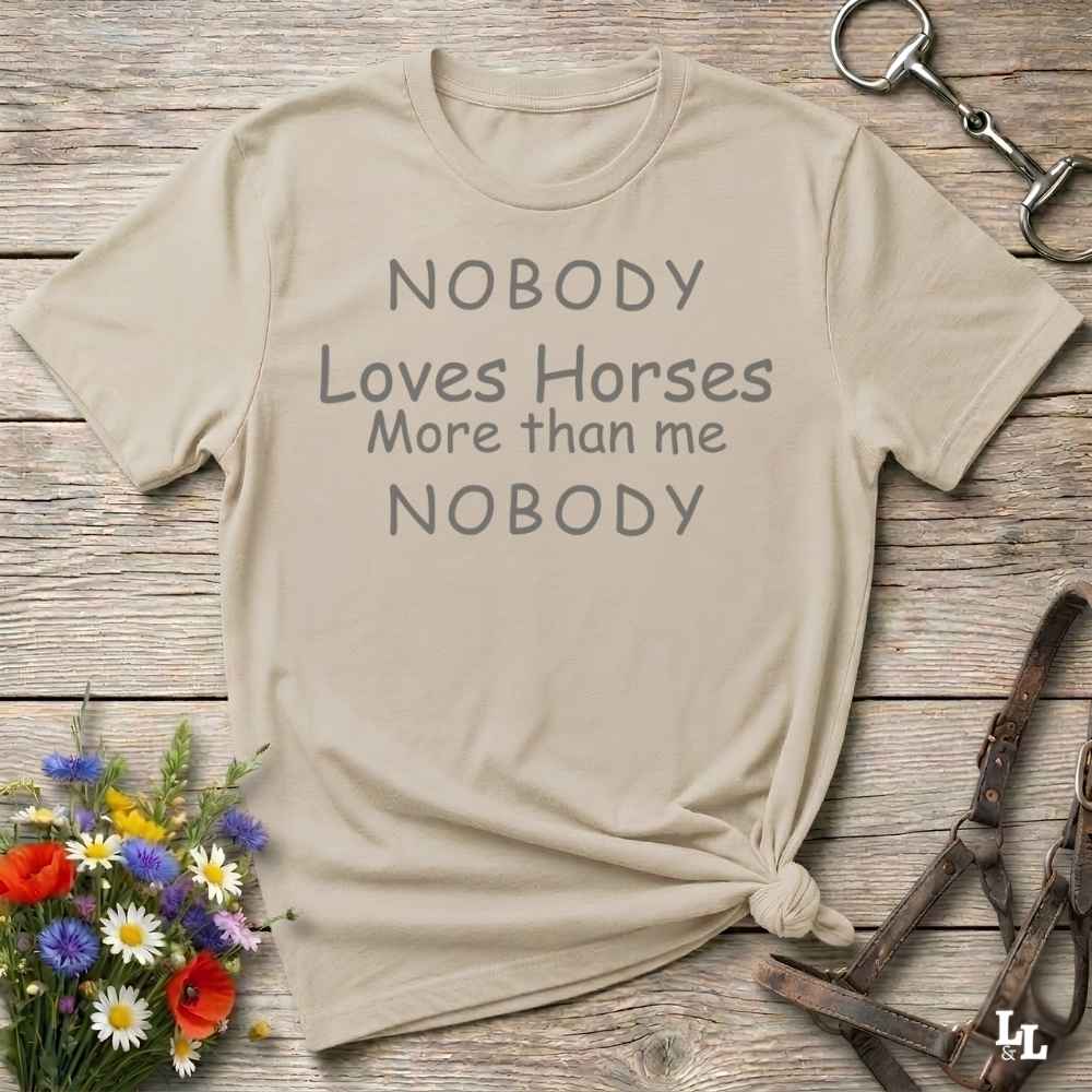 A beige graphic t-shirt with the text 'NOBODY Loves Horses More than me NOBODY' printed in gray, styled in a flat-lay on a rustic wooden background with equestrian accessories and flowers.