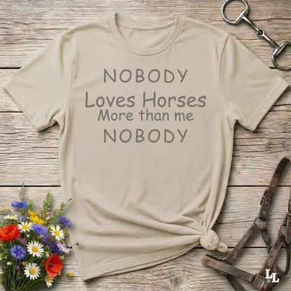 A beige graphic t-shirt with the text 'NOBODY Loves Horses More than me NOBODY' printed in gray, styled in a flat-lay on a rustic wooden background with equestrian accessories and flowers.