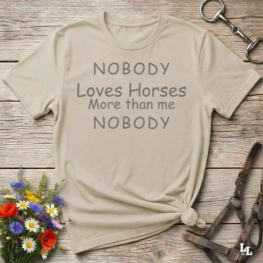 A beige graphic t-shirt with the text 'NOBODY Loves Horses More than me NOBODY' printed in gray, styled in a flat-lay on a rustic wooden background with equestrian accessories and flowers.