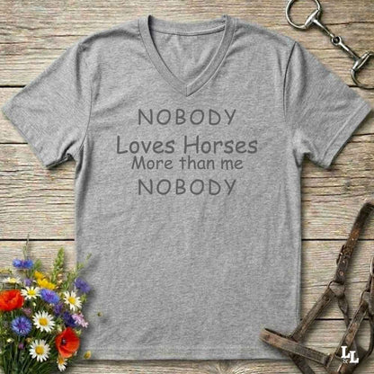 A heather gray V-neck t-shirt with the text 'NOBODY Loves Horses More than me NOBODY' printed in dark gray. The shirt is displayed flat on a rustic wooden background, styled with equestrian accessories (a horse bit in the top right and a halter in the bottom right) and a small bouquet of colorful wildflowers in the bottom left.
