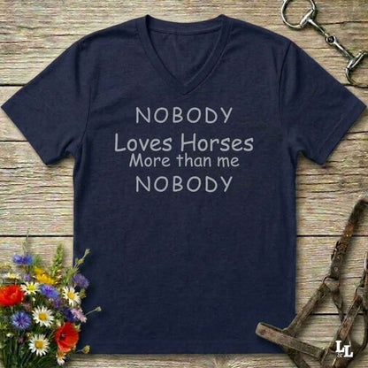 A navy blue V-neck t-shirt with the text 'NOBODY Loves Horses More than me NOBODY' printed in white on the front, laid flat on a rustic wooden background with equestrian-themed props.