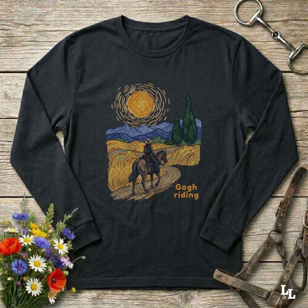 A black long-sleeve t-shirt with a 'Gogh riding' Van Gogh-inspired art print, displayed flat on a rustic wooden background with decorative flowers and horse tack.