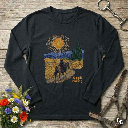 A black long-sleeve t-shirt with a 'Gogh riding' Van Gogh-inspired art print, displayed flat on a rustic wooden background with decorative flowers and horse tack.
