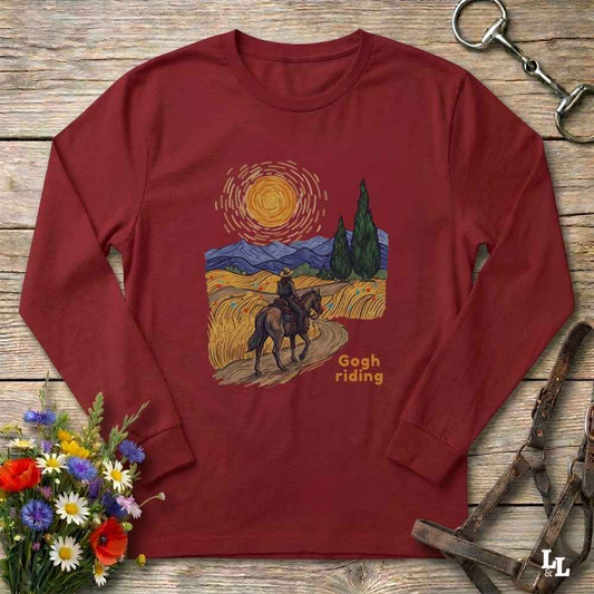 A long-sleeve crewneck t-shirt in maroon red, laid flat on a rustic wooden surface. The shirt features a colorful graphic print in the center depicting a cowboy riding a horse through a stylized, impressionistic landscape with a sun and mountains, captioned 'Gogh riding'.
