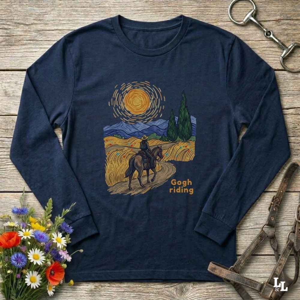 A navy blue long-sleeve t-shirt with a graphic print of a cowboy riding a horse through a field in a Van Gogh-inspired style, with the text 'Gogh riding' printed below. The shirt is displayed flat on a rustic wooden background with equestrian and floral props.