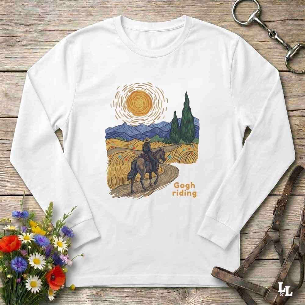 A white long-sleeve t-shirt with a vintage-style Van Gogh-inspired graphic of a rider on horseback in a golden field, laid flat on a rustic wooden surface with decorative flowers and leather tack around it.
