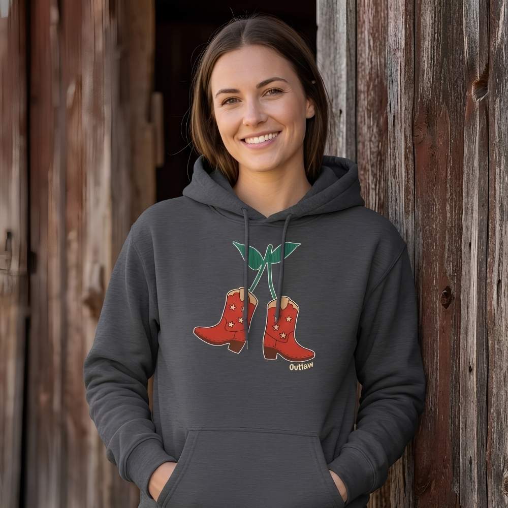 Woman wearing a dark gray hoodie with a graphic design of red boots and a green plant, standing against a wooden barn. Cherry Cowboy Boots Hoodie for Horse Riders, Barn Wear Hoodie, Trail Riding Top, horse girl hoodie, horse mom hoodie, horse lover, horse girl, Funny horse lover gifts, Funny horse hoodies, Horse girl gifts, Equestrian graphic hoodies, fathers day gift, Sarcastic horse hoodie