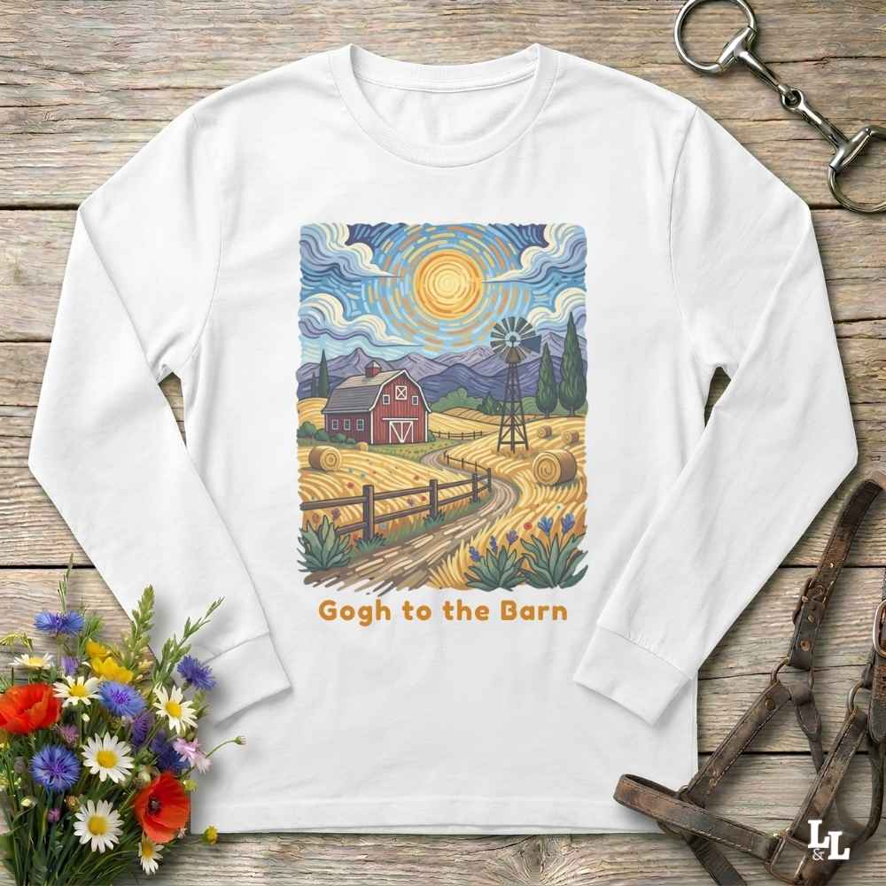 A white long-sleeve t-shirt with a colorful, stylized art print of a farm scene in a Van Gogh-inspired style, featuring a red barn, yellow fields, and a windmill, with the text 'Gogh to the Barn' printed below. The shirt is displayed flat on a rustic wooden background with decorative props including flowers, a horse bit, and a leather bridle.