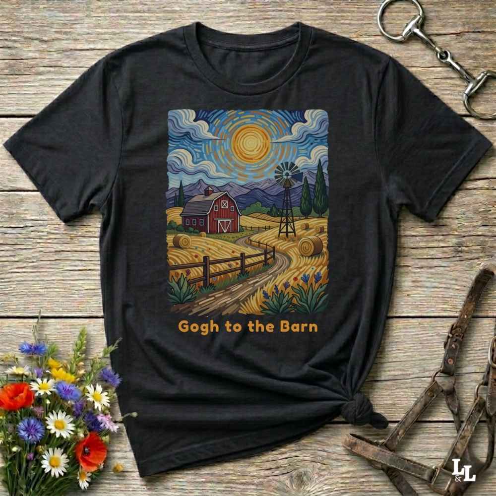 A black graphic t-shirt laid flat on a rustic wooden surface, styled with flowers, a horse bit, and a leather halter. The t-shirt features a colorful art print in a Van Gogh-inspired style showing a red barn, a windmill, and a yellow sun, with the text 'Gogh to the Barn' printed below.