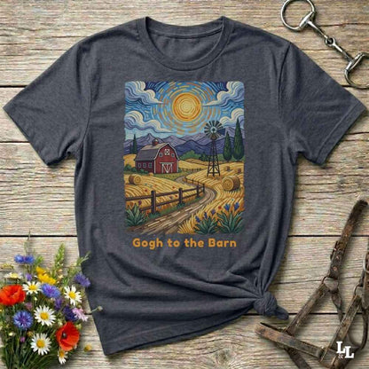 A dark heather gray graphic t-shirt laid flat on a rustic wooden surface, styled with a bouquet of wildflowers, a horse bit, and a leather halter. The t-shirt features a colorful print in a Van Gogh-inspired style showing a red barn, hay bales, a windmill, and a yellow sun, with the text 'Gogh to the Barn' printed below.
