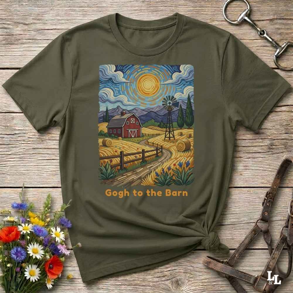 An olive green graphic t-shirt laid flat on a rustic wooden surface, styled with a horse bit, leather straps, and a small bouquet of wildflowers. The t-shirt features a colorful print in the style of Van Gogh's Starry Night depicting a farm scene with a barn, windmill, and the text 'Gogh to the Barn'.