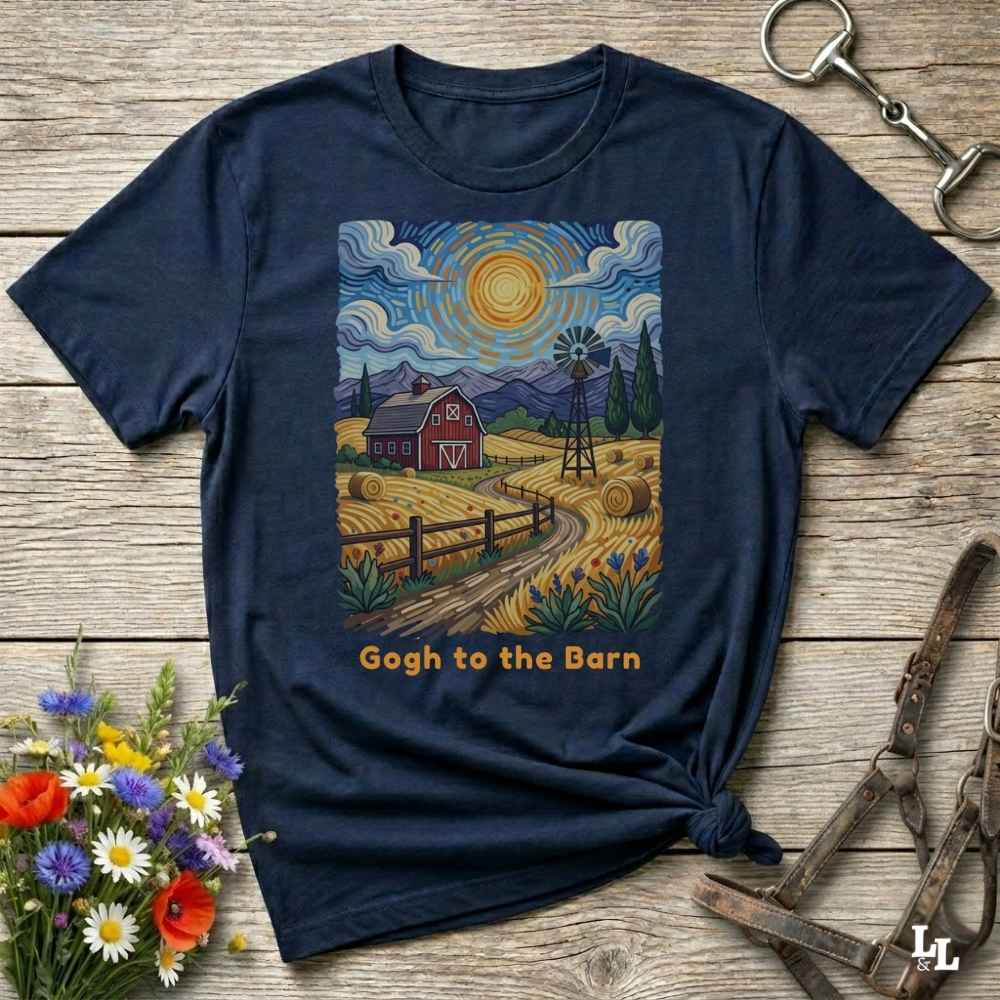 A navy blue graphic t-shirt laid flat on a rustic wooden surface, styled with equestrian tack and a small bouquet of wildflowers. The t-shirt features a colorful print in a Van Gogh-inspired style showing a red barn, windmill, hay, and mountains under a swirling sky, with the text 'Gogh to the Barn' printed below.
