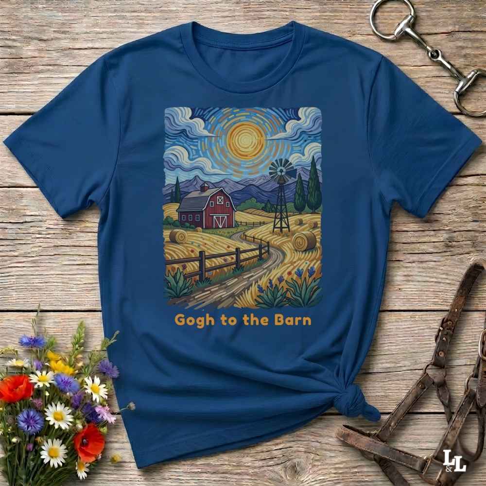 A navy blue graphic t-shirt laid flat on a rustic wooden surface, styled with a bouquet of flowers, a horse bit, and a leather halter. The t-shirt features a colorful print in a Van Gogh-inspired style showing a red barn, windmill, and haystacks, with the text 'Gogh to the Barn' printed below.