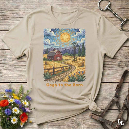 A beige graphic t-shirt laid flat on a rustic wooden surface, styled with a small bouquet of flowers, a metal clasp, and leather straps. The t-shirt features a colorful illustration of a farm scene with a barn, windmill, and haystacks, with the text 'Gogh to the Barn' printed below.