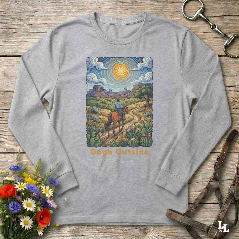 A light gray long-sleeve t-shirt with a colorful graphic print of a cowboy riding a horse along a desert path under a bright sun, with the text 'Gogh Outside' below. The shirt is displayed flat on a rustic wooden background with decorative props including a bouquet of flowers, a horse bit, and a leather strap.