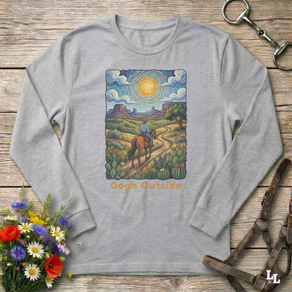 A light gray long-sleeve t-shirt with a colorful graphic print of a cowboy riding a horse along a desert path under a bright sun, with the text 'Gogh Outside' below. The shirt is displayed flat on a rustic wooden background with decorative props including a bouquet of flowers, a horse bit, and a leather strap.