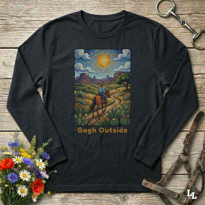 A black long-sleeve t-shirt with a colorful graphic print of a cowboy riding a horse through a desert landscape under a swirling sky, with the text 'Gogh Outside' below. The shirt is displayed flat on a rustic wooden background with decorative flowers and horse tack.