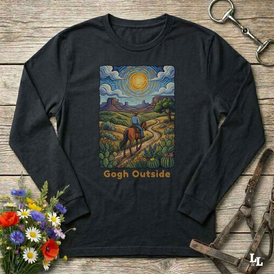 A black long-sleeve t-shirt with a colorful graphic print of a cowboy riding a horse through a desert landscape under a swirling sky, with the text 'Gogh Outside' below. The shirt is displayed flat on a rustic wooden background with decorative flowers and horse tack.