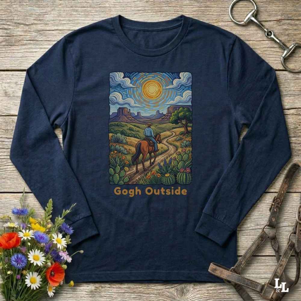 A navy blue long-sleeve t-shirt with a colorful graphic print of a cowboy riding a horse along a desert path under a bright sun, with the text 'Gogh Outside' below. The shirt is displayed flat on a rustic wooden background with decorative props including flowers, a horse bit, and a leather halter.