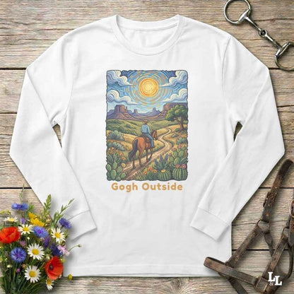 A white long-sleeve t-shirt with a colorful graphic print of a cowboy riding a horse along a desert path under a bright sun, with the text 'Gogh Outside' below. The shirt is displayed flat on a rustic wooden background with decorative equestrian and floral props.
