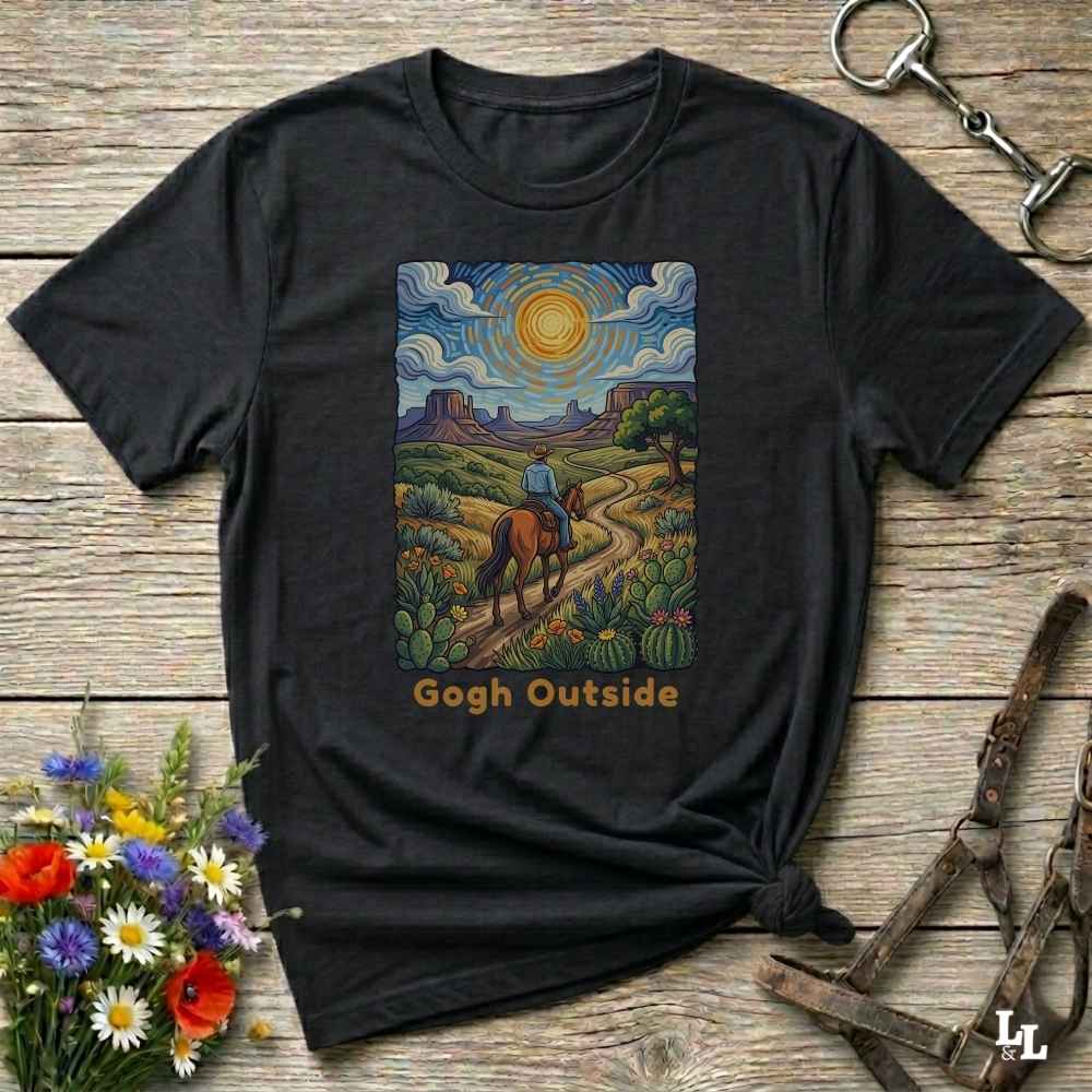 A black graphic t-shirt laid flat on a rustic wooden surface, styled with a small bouquet of flowers, a horse bit, and a leather halter. The t-shirt features a colorful central print depicting a desert landscape with a rider on a horse under a swirling sky, with the text 'Gogh Outside' printed below.